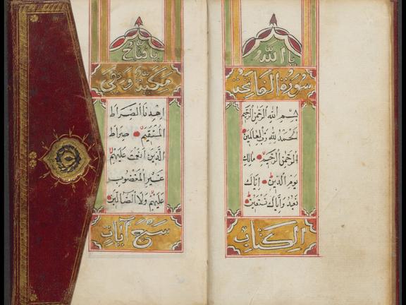 Das Bild zeigt den aufgeschlagenen Koran, dessen Seiten mit einem gelb, grün, rotem Muster verziert sind. An der linken Seite ist der Klappdeckel zu sehen, welcher mit einem rot, golden gestaltet ist.